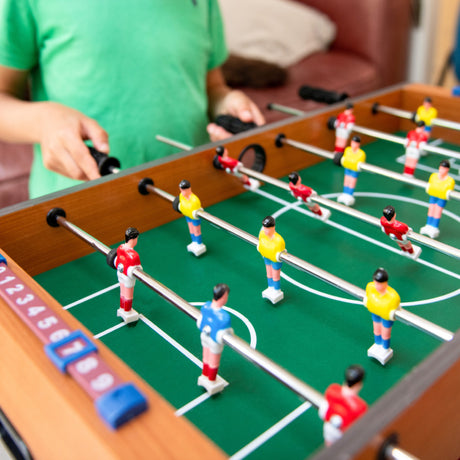 Tabletop foosball game with children playing, featuring miniature soccer players in yellow and red uniforms on a green field, perfect for home entertainment or kids' gaming rooms.